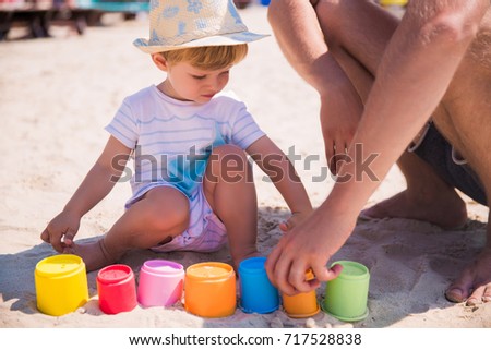 The little cute blonde boy playing on sand on the beach with plastic color buckets and studying colors