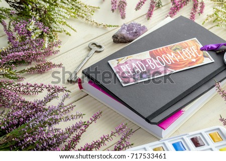 Book with bookmark, heather branches, and a box of watercolor paints on a white wooden table. Royalty-Free Stock Photo #717533461