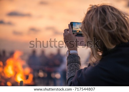 Woman is taking picture of sunset with smart phone. Romantic bonfire night at seaside. People gathering together to celebrate Night of ancient lights. Large burning campfire with soft glowing flame.