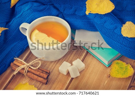 White cup with hot tea and lemon, books and knitted scarf or sweater on wooden table. Autumn leafs