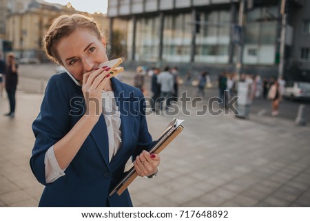 Busy woman is in a hurry, she does not have time, she is going to eat snack on the go. Worker eating and talking on the phone . Businesswoman doing multiple tasks.   Business female person.
