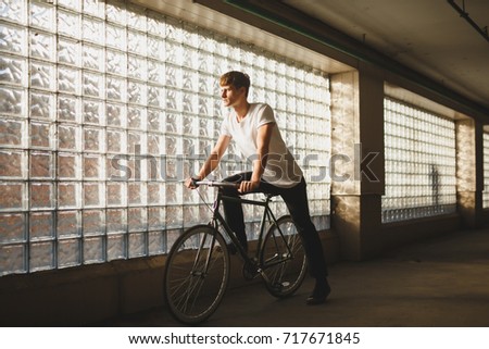 Portrait of cool boy riding classic bicycle while dreamily looking aside. Young thoughtful man in white t-shirt standing with bicycle with glass wall on background