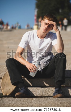 Portrait of boy with brown hair sitting on stairs and holding bottle of water while tiredly looking aside. Photo of young thoughtful man in white t-shirt sitting with skateboard nearby