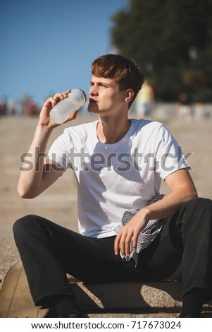 Cool boy with brown hair sitting on stairs and drinking water while dreamily looking aside. Photo of young thoughtful man in white t-shirt sitting and holding bottle of water with skateboard nearby