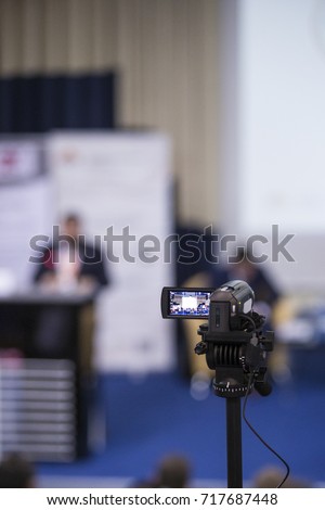 Back View of Compact Videocamera. Positioned Against Blurred Background with Host Speaking on stage. Vertical Image