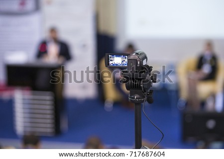 Back View of Compact Videocamera. Positioned Against Blurred Background with Host Speaking on stage. Horizontal Image