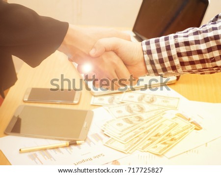 The business worker making handshake ,in blurry light design,among investment chart paper laptop mobile phone and Us Dollars ,in success work concept,merger and acquisition concepts,Vintage light tone