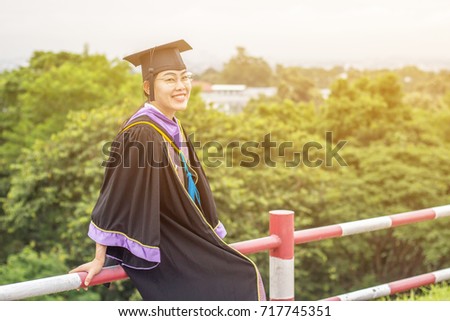 The Asian woman wearing black graduate dress and hat with colorful stripes is being taken photo on the asphalt road in the outdoor park with the trees background and flare from the picture edge.