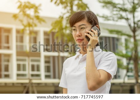 The Asian woman in white T shirt is talking to somebody by mobile phone at the outside with the group od business building and trees with the flare from the picture corner. 