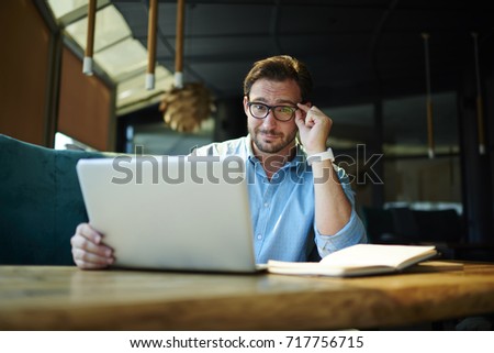 Handsome marketing expert looking through eyeglasses skeptically while browsing web pages with advertising content portrait of professional editor doubting in publication while proofreading on laptop
