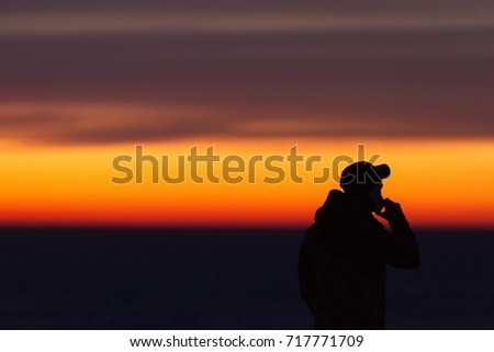 Silhouette of the man with mobile/cellphone during sunset at the seaside. Man in a baseball cap.