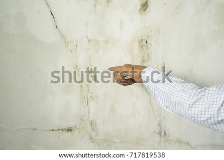 Left hand of young Asian man wearing white long sleeve shirt,  pointing at dirty mold and fungus on building white cement wall texture background