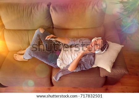 Teen lying face up on couch with headphones and smartphone consuming multimedia content top view