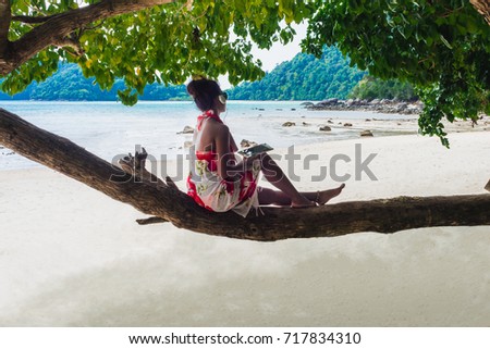 Happy woman in bikini relaxing under tree near the beach listening to music on headphone