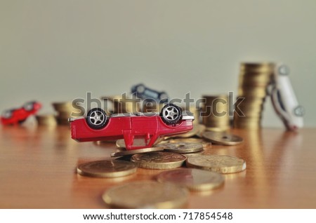 Debt concept, miniature red truck overturn upside down on pile of money and blur toy car on rolls ladder of coins on wood table in low light 