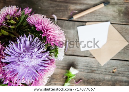 Top view Multicolor aster flowers bouquet, pen, blank white greeting card and craft paper envelope on rustic wooden table.. Postcard mock up. Autumn flowers. Selective focus. Space for text. Flat lay.