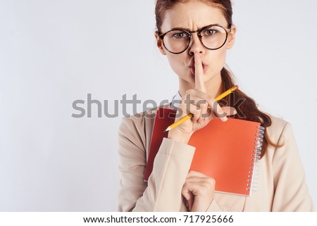 young teacher with glasses looking at camera holding finger on lips in hand pencil and orange notebook on light background portrait                               