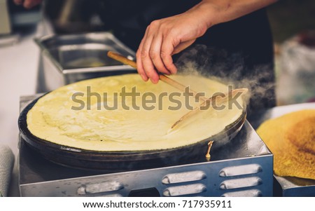 Making of crepes pancakes in open market festival fair. A hand is making crepes outdoors on a metal griddle with wooden stick on a outdoor summer fest. Royalty-Free Stock Photo #717935911