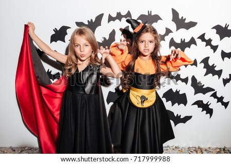 Couple of two scary little girls dressed in halloween costumes posing with arms raised and bats on a background
