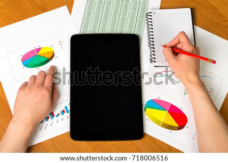 Tablet, woman's hand holding a pencil, notebook, sheets of paper account and charts on the desktop background.