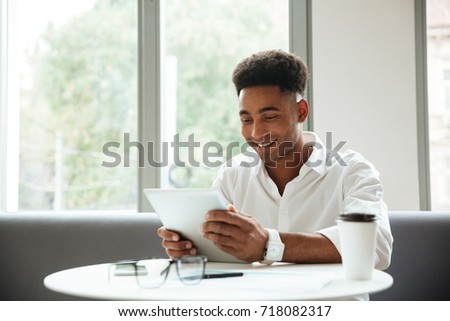 Image of cheerful young african man sitting coworking by tablet computer. Looking aside.