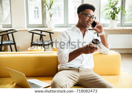 Picture of handsome young african man sitting coworking by laptop computer. Looking aside talking by phone drinking coffee.