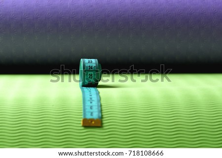 Measuring tape roll in cyan color on green and purple texture background. Workout and sport concept. Flexible ruler in blue lying on yoga mat, defocused. Shaping and fitness equipment, copy space