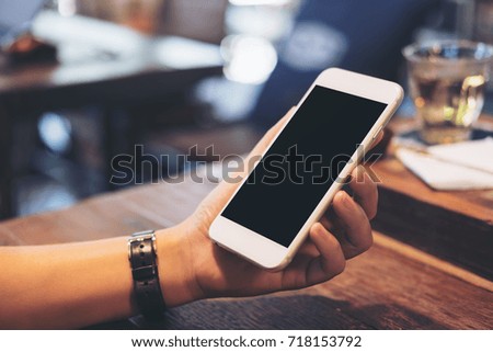 Mockup image of a woman's hands holding white mobile phone with blank black screen with tea cup on wooden table in vintage cafe