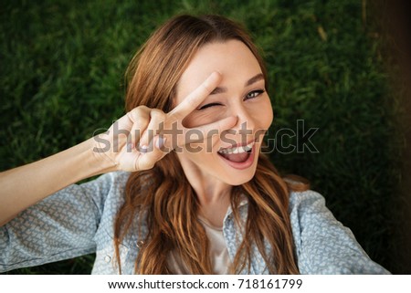 Close up top view of happy brunette woman lying on grass in park and making selfie while showing peace sign