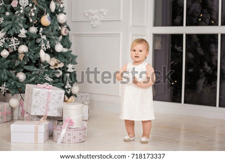 Little girl child is standing in a white dress near the Christmas tree, happy holiday new year with toys,  
gifts, flashlight, waiting for a miracle. bright room, modern interior