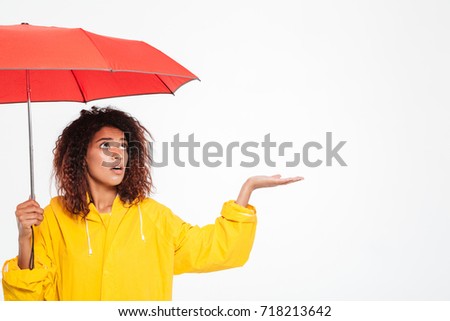 Picture of confused african woman in raincoat hiding under umbrella and waiting rain over white background