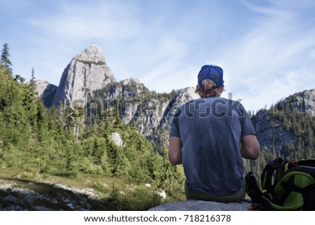 Adventurous young man is sitting and relaxing on the rock while enjoying the beautiful scenery. Picture taken on the ascent to Skypilot Mountain near Squamish, North of Vancouver, BC, Canada.