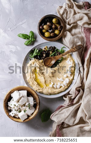 Homemade traditional spread hummus with chopping olives, oil and herbs on blue plate, served with olives, feta cheese, spoon, textile on gray texture background. Mediterranean snack. Flat lay