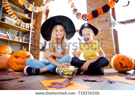 Treat or trick! dessert! Cheerful amazed excited small kids in carnival head wear, with colorful treats, carved pumpkins, fall leaves, orange papers near them, sit on the brown wooden floor at home