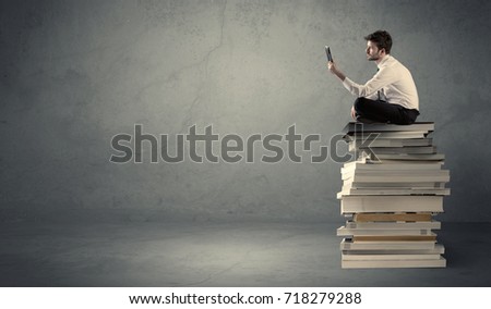 A serious businessman with laptop tablet in elegant suit sitting on a stack of books in front of dark grey background