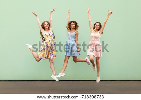 Three happy excited young adult women in casual dresses celebrate victory and jumping over green background. Outdoor