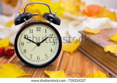 Old vintage alarm clock and old book with autumn yellow leaves on a wooden table.