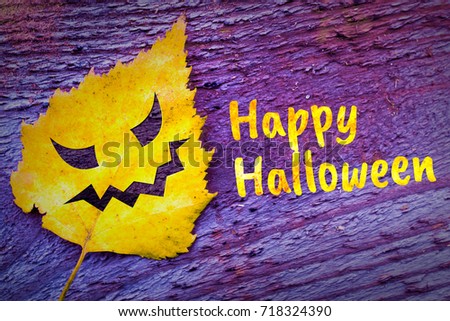 Halloween face on a yellow autumn leaf on wooden background. With Happy halloween inscription