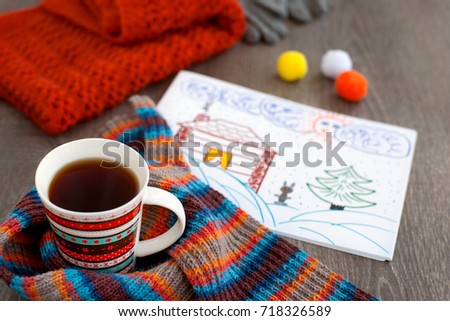 A mug of tea and a picture about winter. A cup of tea, a knitted scarf and a picture of snow, a Christmas tree and a warm house remind us of the winter. Tea for warming in the cold season.