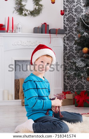 Cute happy boy in santa hat holding toy. Christmas present on holiday morning in beautiful room. Child got Xmas gift near decorated fir tree and fireplace posing on camera. Winter holidays concept