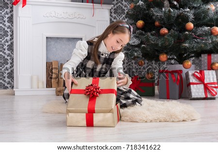 Beautiful happy girl unwrap christmas present box on holiday morning in beautiful room interior. Female child open Xmas gift near decorated fir tree and fireplace. Winter holidays concept