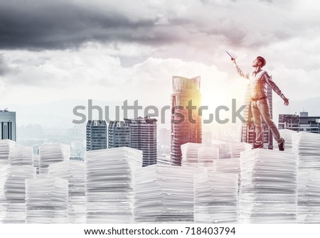 Side view of man in casual wear keeping hand with book up while standing on pile of documents with cityscape and sunlight on background. Mixed media.