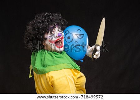 Angry ugly clown wants to kill a balloon in a cap on a black background