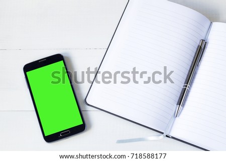 Smart phone with green screen and open notepad on desk. Cellphone concept background. Copy space