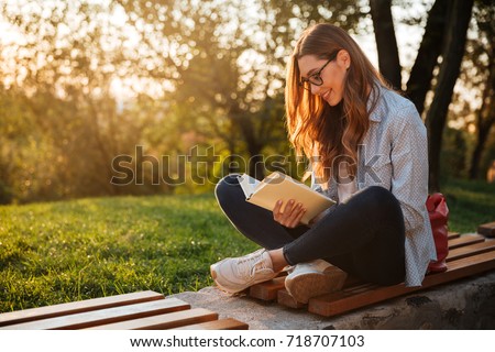 Side view of pleased brunette woman in eyeglasses sitting on bench and reading book in park Royalty-Free Stock Photo #718707103
