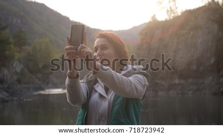 Young woman is taking pictures on the phone against the background of big mountains and the green mountain river. selfie or self-portrait on a smartphone