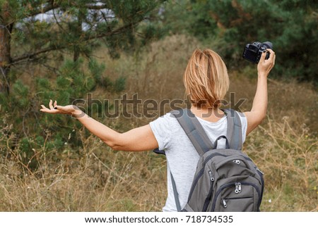 Young lady walking on at forest with digital camera. Hiker with backpack taking pictures of the summer landscape outdoors in nature. Healthy active lifestyle