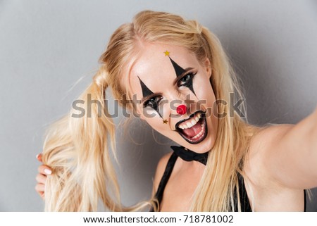 Close up portrait of a crazy blonde woman in bright halloween clown make-up taking a selfie isolated over gray background