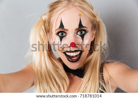 Close up portrait of an excited blonde woman in bright halloween clown make-up taking a selfie isolated over gray background