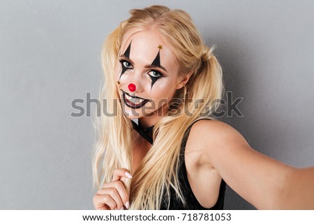 Close up portrait of a happy young blonde woman in bright halloween clown make-up taking a selfie isolated over gray background